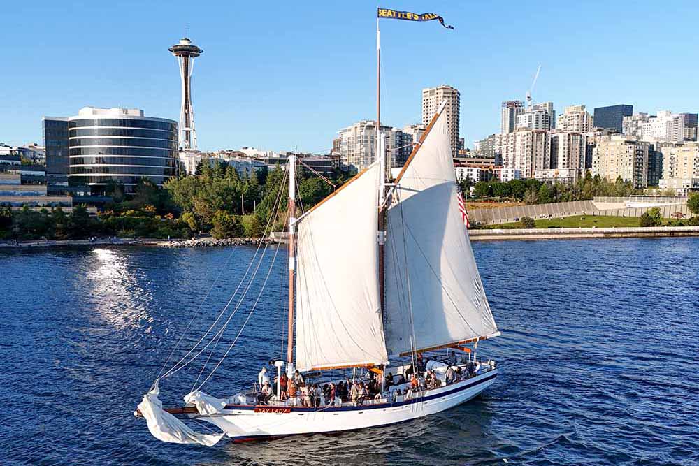 What to do between World Cup matches in Seattle. The Bay Lady tall ship sailing under full canvas on Elliott Bay with Waterfront Park and the Great Wheel visible along the Seattle waterfront on a clear sunny day.
