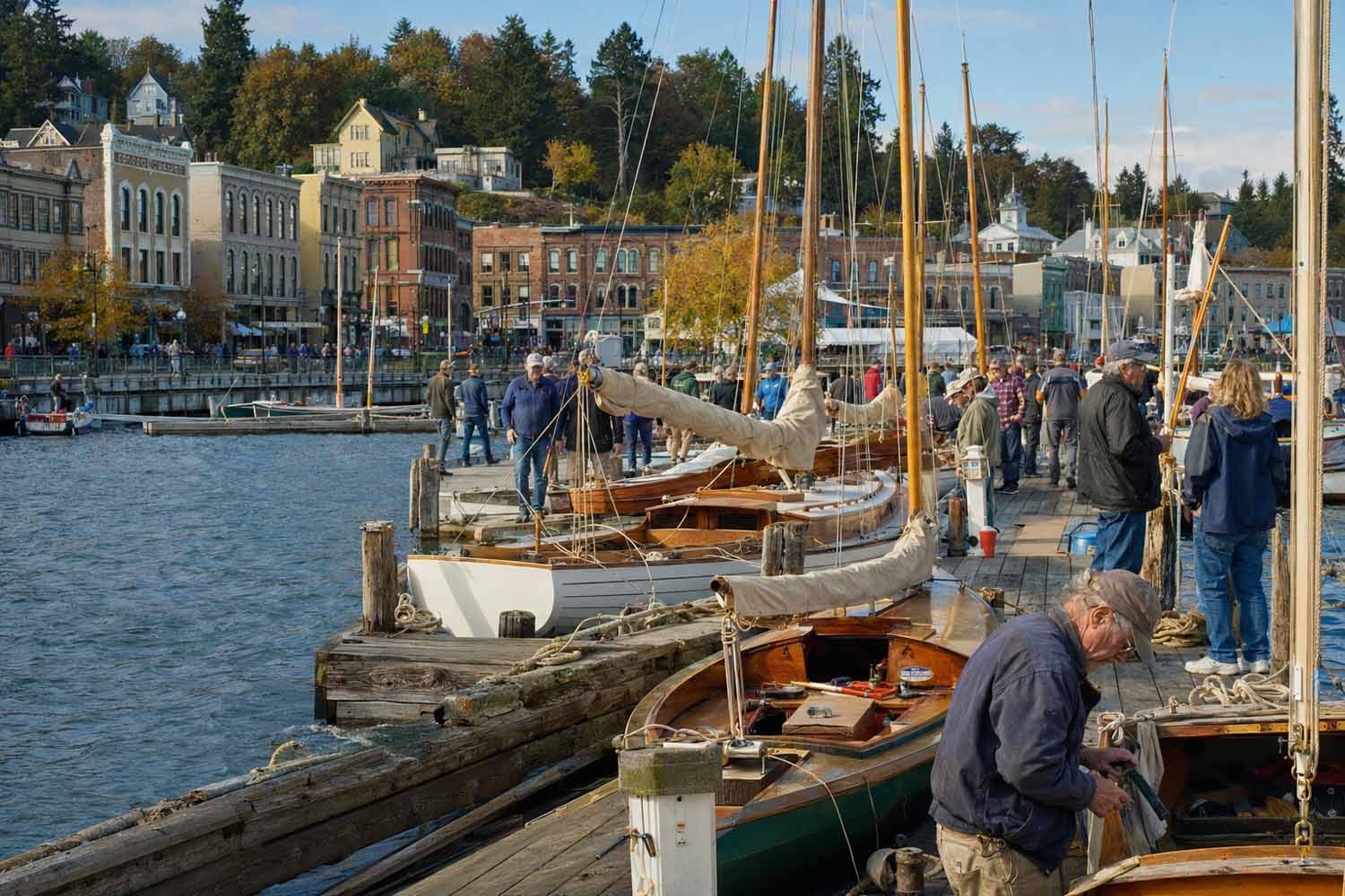 19th-century Puget Sound shipping. Wooden sailboats line a Port Townsend dock as visitors gather beside preserved Victorian waterfront buildings.